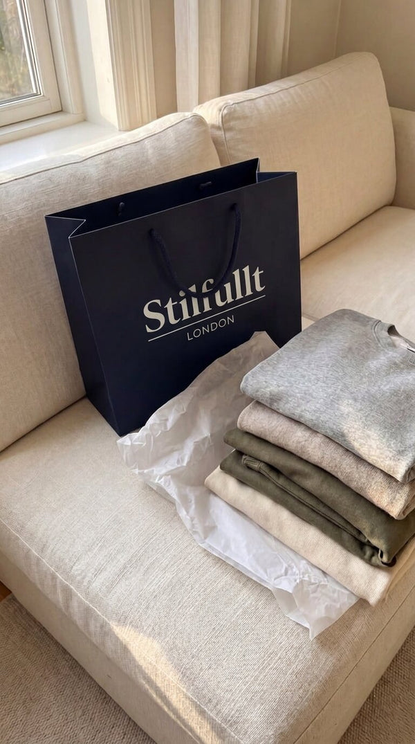 Stilfullt shopping bag beside a stack of folded knitwear on a cream sofa