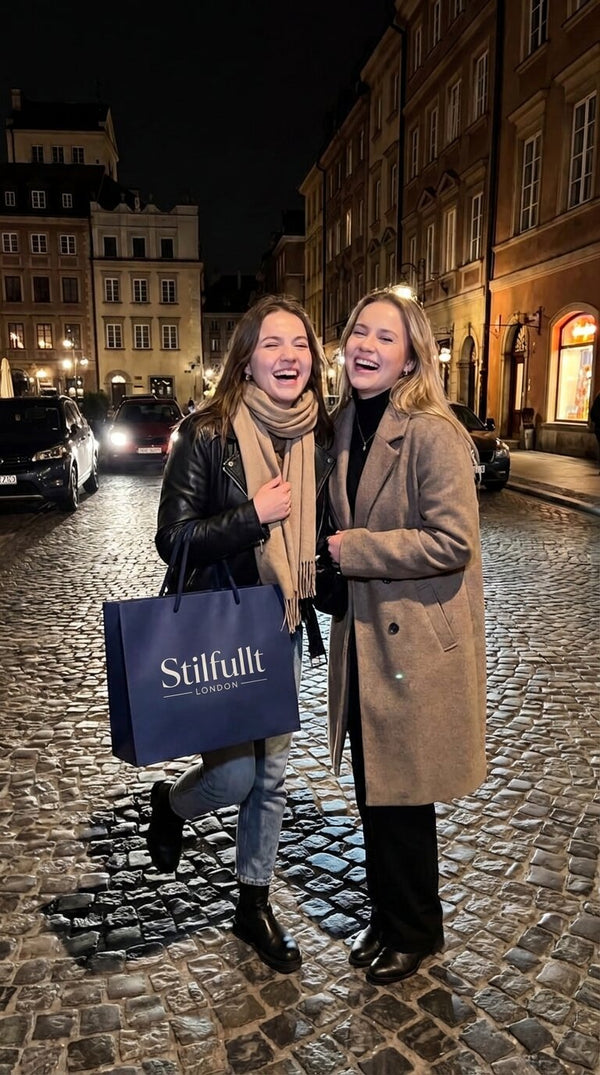 Two friends laughing in winter coats on a cobbled street at night, one holding a Stilfullt shopping bag